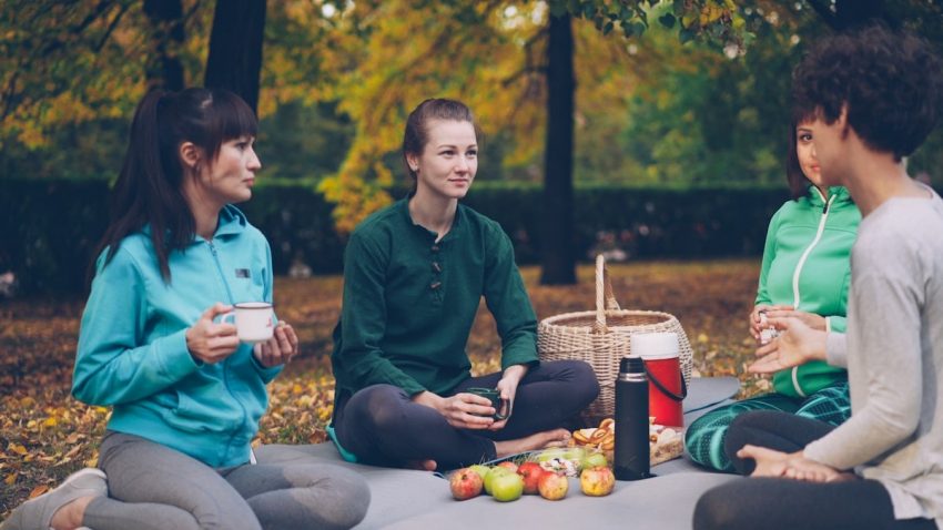 Cheerful young women friends are resting on mats having picnic talking and drinking tea after yoga class outdoors on beautiful autumn day. Food and basket are visible.
